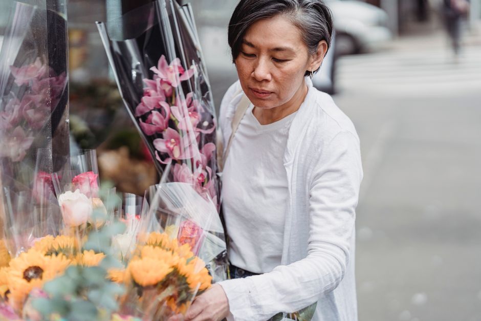 A mature woman with short dark hair, wearing a white top and light-colored cardigan, stands outdoors on a grey paved street, carefully examining a display of vibrant floral bouquets outside a flower s
