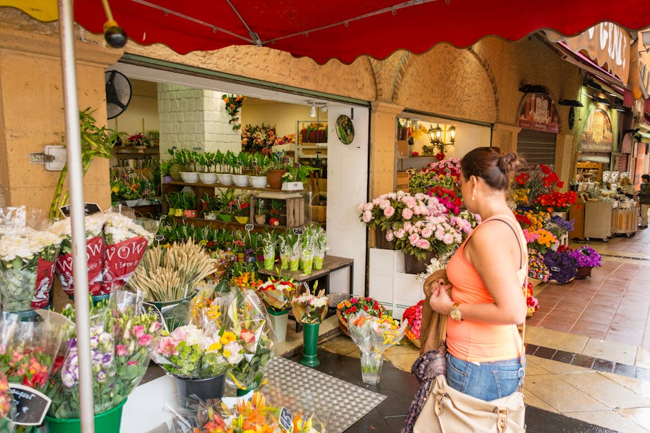 A woman in a peach sleeveless top and blue jeans is holding a bouquet of red and pink flowers while standing outside a flower shop on Yiewsley High Street. The shop, part of flowersyiewsley.co.uk, dis