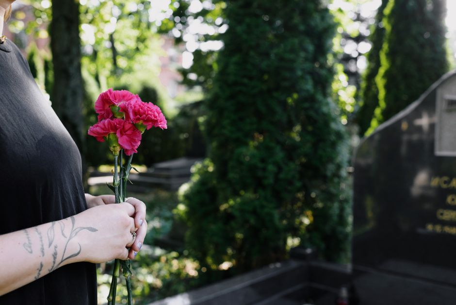 A person's hands, with visible tattoos on the wrist and arm, are gently holding a small bouquet of vibrant pink carnations with ruffled petals and green stems. The bouquet is wrapped in clear plastic,