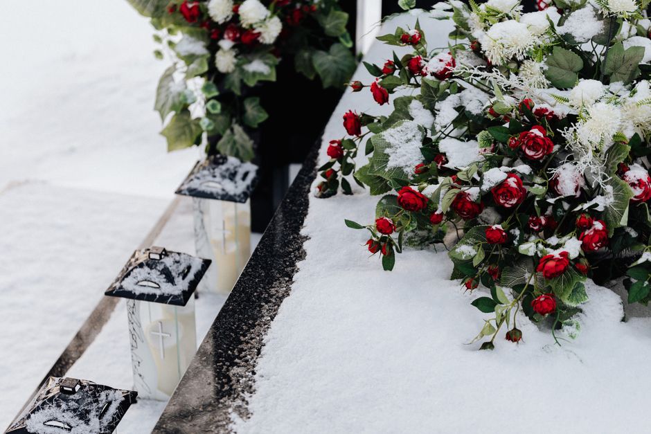 A floral arrangement of red roses, white flowers, and green foliage, partially dusted with snow, rests on a snow-covered surface at a cemetery. The bouquet is placed on a metal ledge alongside three w