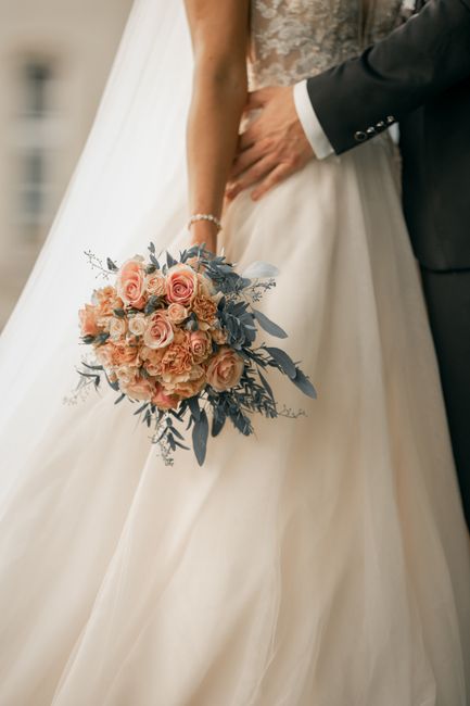 A close-up of a bride holding a lush bouquet of pale pink roses, peach-colored ranunculus, and soft blue-gray eucalyptus leaves, accented with delicate baby's breath. The bouquet has a natural, slight