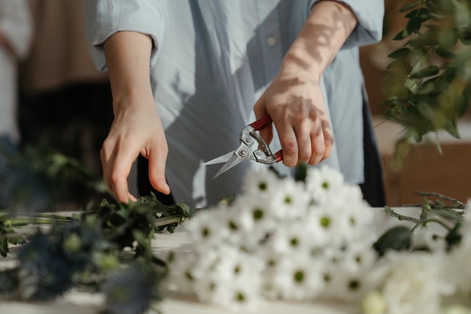 A person wearing a light grey shirt is using a small pair of red-handled scissors to trim stems of white daisies with dark green centers, arranged in a bouquet on a white table. Around the bouquet are