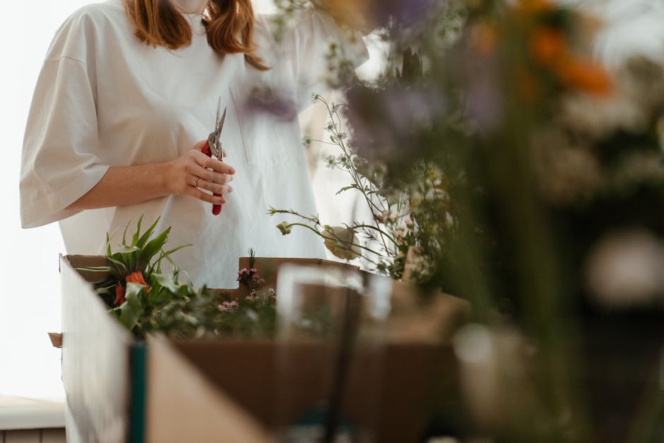 A professional florist, dressed in a white apron, is working at a wooden table arranging fresh flowers for a floral gift or bouquet arrangement. The florist is holding a pair of pruning scissors in on