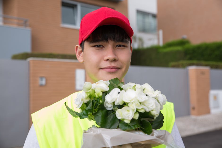 A young delivery person, wearing a red cap and a high-visibility yellow vest, is holding a bouquet of fresh white flowers wrapped in white paper. The bouquet features tightly clustered white roses or