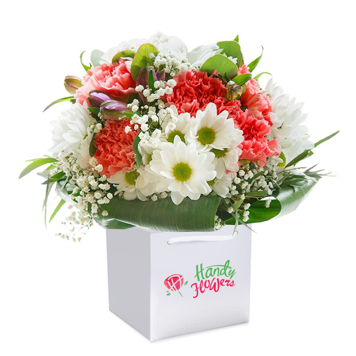 Standing proudly in a square white gift bag, this luxurious bouquet is a celebration of fresh, local floristry in Yiewsley. The piece is shaped into a full, rounded form, filled with the ruffled, abundant petals of coral-pink and peach carnations. Their soft, romantic tones combine with darting, bright white daisies whose lime-green centers glisten invitingly, recalling springtime walks near Yiewsley Recreation Ground. Nestled throughout the arrangement, a scattering of delicate baby's breath adds a drifting, cloudlike softness, set against deep green foliage and broad, glossy leaves that embrace the base. To the side, only just visible, are a few tightly closed, violet-tipped buds, their muted shades suggesting grace and quiet promise. The bouquet's presentation is as crisp as its design-set against a pure white background that draws attention to every nuance of shade, texture, and line. The gift bag, with its neat white handles, hints at convenience and care, ready for local delivery. Whether marking a milestone birthday, an anniversary, or simply spreading joy in Yiewsley and nearby Hayes, this bouquet is an ideal choice for those seeking freshness and heartfelt floral artistry delivered straight from a neighbourhood expert.