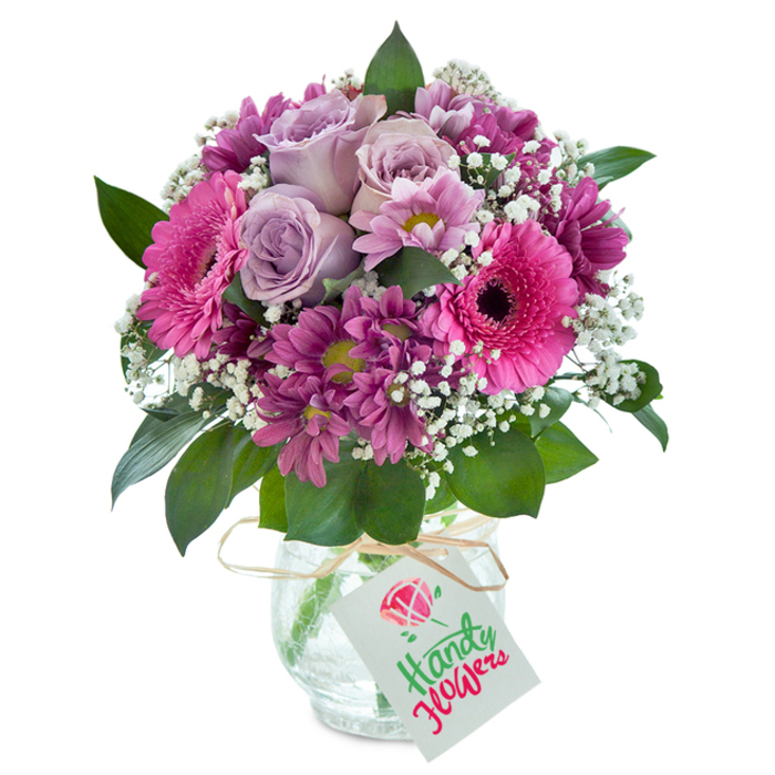 Taking centre stage in the bright, inviting light typical of a Yiewsley flower shop, this opulent bouquet is a celebration of pink, purple, and rich lavender hues. At the heart, soft lavender roses gently unfurl, inviting a sense of calm elegance familiar to those strolling the leafy avenues of Harmondsworth Road. Surrounding the roses, bold hot pink Gerbera daisies radiate cheer, their dark-eyed centers anchoring the arrangement, while clusters of deep magenta chrysanthemums weave texture and intrigue throughout the dense composition. Wispy white baby's breath is threaded liberally between the blooms, its dainty blossoms catching the light and evoking the gentle airiness of a spring morning near Little Britain Lake. Lush, verdant leaves peek out from beneath the profusion of flowers, grounding the arrangement and enhancing the bouquet's visual harmony. The arrangement is cradled in a clear glass vase with an intricate crackled effect, a tactile detail that invites the eye and hand alike. A band of natural raffia ties a light, rustic note around the neck, from which a neat white tag dangles-ideal for inscribing a birthday wish, anniversary note, or words of gratitude. This bouquet, thoughtfully designed for delivery across Yiewsley and West Drayton, expresses care and celebration in every petal, embodying the skilled touch and community spirit of local florists.