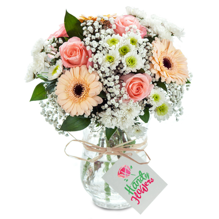 A delicate arrangement sits proudly on a luminous white field-a bouquet that touches on the best of Yiewsley's local floral artistry. Focal to the design are peach-pink roses, their elegantly layered petals curling outwards, celebrated for their classic beauty and subtle scent. Alongside rise two peach and light orange gerbera daisies, open-faced with striking dark cores, lending both energy and calm. Cloudlike tufts of white gypsophila wind between the key blooms, enhancing a sense of airiness and lightness, while clusters of snowy chrysanthemums, some cradling vivid green hearts, bring volume and lively contrast reminiscent of springtime along the canal. Sprigs of fresh eucalyptus peek out, a nod to the district's lush pockets of greenery. The composition is presented in a textured glass vase with glimmering water visible beneath neat, trimmed stems-its neck adorned with a simple rustic twine and a white, blossom-emblazoned tag. This bouquet's soothing palette and artisanal touch make it perfect for birthdays, anniversaries, or new beginnings in Yiewsley, whether destined for a home on Fairfield Road or a confetti-decked family occasion.