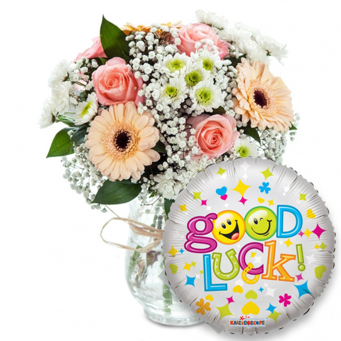 Framed against a bright white background, this uplifting 'Good Luck!' gift combines a softly romantic bouquet with a festive foil balloon. The floral design, placed upper left and centre, gathers soft peach and blush-pink roses whose layered petals suggest a gentle scent, paired with peachy-orange Gerbera daisies that carry darker brown centres for a striking contrast. Faint, airy sprays of white baby's breath spill through the arrangement like delicate lace, while clusters of white and lime-green chrysanthemum florets punctuate the composition with crisp, energetic form. Rich green foliage slips between the blooms, adding structure and a shadowed depth that makes the pastel tones sing. The simple clear glass vase shows the stems and is finished with a rustic twine bow-a handcrafted touch recalling local markets and artisan stalls near Yiewsley High Street and the small community green.
On the right, overlapping the bouquet's lower edge, a shiny silver Mylar balloon floats; its surface is alive with small colourful shapes-blue and pink hearts, yellow and blue stars, tiny orange and green cross-like motifs-and proclaims Good Luck! in a bubbly, multi-coloured type. The word Good uses playful purple letters with the two 'o's replaced by yellow smiley faces-one winking, the other grinning with its tongue out-while Luck follows in green, blue, orange and yellow tones, finished with a bright exclamation mark. A discreet KALEIDOSCOPE logo sits at the bottom of the balloon. The image conveys a quiet optimism perfect for send-offs, graduations, or any new beginning in the Yiewsley and Stockley Park community.