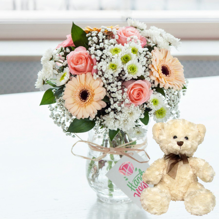 A serene and affectionate still life is arranged on a bright white table, capturing the mood of a thoughtful anniversary surprise ready to be sent to a loved one in Yiewsley. Dominating the scene is a compact, rounded bouquet displayed in a clear glass jar-style vase, allowing you to glimpse the pale green stems gathered neatly inside. The flowers themselves form a soft, pastel harmony: blush pink roses opening in gentle spirals; peach-toned gerbera daisies with distinctive dark brown centres radiating from the middle; and generous touches of delicate white baby's breath that seem to float in between. Interspersed are clusters of white chrysanthemums with subtle lime-green hearts, their many fine petals giving texture and fullness to the arrangement. Around the neck of the vase, a simple piece of natural twine has been tied with care, knotted into a small bow and supporting a white rectangular gift tag. On the tag, a stylised pink diamond or heart-shaped icon appears above flowing green script that hints at words such as handmade or with love, emphasising a personal, crafted feel. To the right of the flowers sits a cream-coloured teddy bear, plush and inviting, positioned upright as if keeping watch over the bouquet. Its fur looks soft and lightly tousled, with dark bead eyes, a small brown nose, and a subtle stitched smile that conveys comfort and nostalgia, complemented by a neat brown satin ribbon around its neck. Soft, natural light floods the scene from a blurred background, perhaps suggesting a window overlooking Yiewsley's calm streets, creating gentle reflections on the white surface and enhancing every pastel shade. The overall composition radiates tenderness and quiet celebration, ideal for marking anniversaries, new arrivals, or any heartfelt occasion where flowers and a cuddly companion say more than words.