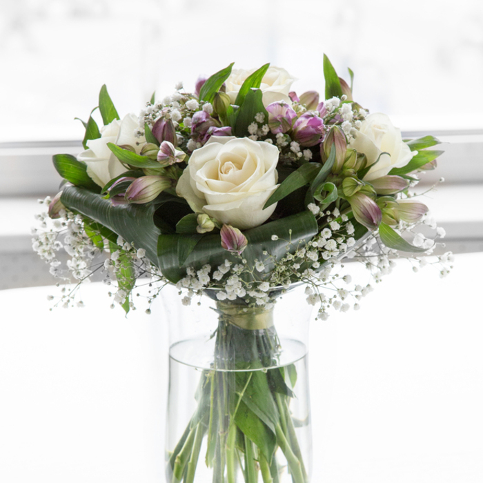 A refined, comforting bouquet dominates the frame, poised in a clear cylindrical glass vase that lets every straight green stem show through the clean water. The arrangement is composed in soft whites and cool purples, reminiscent of a quiet walk through Yiewsley's tree-lined streets on a bright but reflective afternoon. At its core, several white roses open in gentle spirals, their petals thick, velvety, and perfectly shaped, suggesting tenderness and dignity. Interwoven among these are graceful alstroemeria stems, each bloom painted with delicate strokes of lavender, violet, and light green, offering a subdued wash of colour that gently lifts the arrangement without disturbing its calm, respectful mood. Tiny clusters of white baby's breath hover between the larger flowers, like delicate lace or a fine morning mist, filling any empty space with airiness and a sense of softness. A generous collar of foliage surrounds the blooms, from broad, shiny leaves that catch the light to slender, arching greens that add height and structure, evoking the freshness of a well-kept garden near the High Street. The bouquet stands against a bright, almost glowing white background, the light diffused so that no harsh shadows appear-only a serene, even illumination that highlights every petal and leaf. The overall impression is one of stillness, purity, and quiet strength, making this design fitting for sympathy occasions, memorials, or moments of personal remembrance in Yiewsley, when a simple yet elegant floral piece can gently honour a life and offer solace to those who gather.