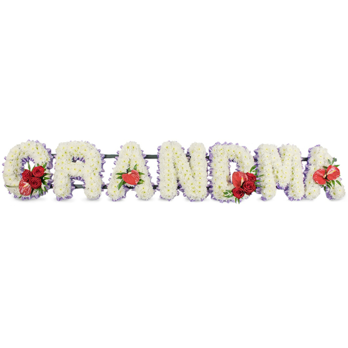 The image shows a dignified floral letter tribute in which the word GRANDMA is spelled out in tall, even block capitals, displayed in a clean horizontal line against a simple white background so the shapes and colours stand out. Each letter is meticulously formed from a blanket of white cushion chrysanthemums or similar small-headed blooms, packed closely together to create a smooth, velvety texture that feels peaceful and comforting. Around the outer edge of every letter runs a slender border of soft mauve or lilac ribbon, just visible beneath the petals, adding a gentle wash of colour and a sense of definition without overpowering the purity of the white flowers. Bright red accents punctuate the calm design at key points. On the leading G, a lush cluster of red roses nestles at the base, alongside a distinctive red-and-white variegated bloom shaped rather like a calla or tulip, giving the starting letter a focal point. The first A features a single, heart-shaped red anthurium, its glossy surface catching the light and echoing the theme of love. The central M is adorned with more red roses and another red anthurium, creating a rich highlight in the middle of the tribute, while the final A is finished with a solitary anthurium, bringing a quiet balance to the composition. The letters are mounted on a dark green framework, likely a mossed or foam-based stand, which provides structure and keeps the arrangement slightly raised. This is very much the sort of bespoke funeral lettering families around Yiewsley might choose when they want flowers to say Grandma in a clear, affectionate way at a local service or graveside committal, offering a lasting symbol of respect and remembrance.
