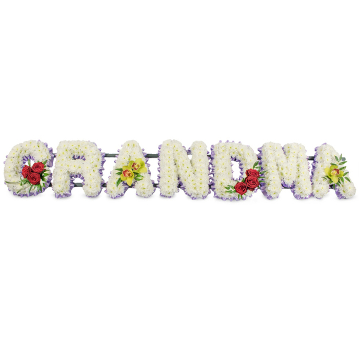 A lovingly crafted sympathy flower tribute spells out "GRANDMA" in large, ornate block letters, laid out in a straight line against a clean, bright white backdrop that makes every petal stand out. Each letter is entirely covered with small, tightly clustered white blooms, likely chrysanthemums, arranged so densely that they create a smooth, cushion-like surface, almost like a soft white blanket of flowers draped across the frame. Beneath this snowy layer, a band of delicate light lavender blossoms peeks out along the base of the letters, giving a gentle purple halo that adds depth and structure without overwhelming the calm, neutral palette. Subtle, meaningful accents personalise the tribute. On the lower left curve of the "G," a cluster of three deep red roses is cradled by dark green leaves, their saturated colour a quiet symbol of passion, respect, and family love. The first "A" from the left features two elegant cymbidium orchids in a fresh yellow-green tone, their throats tinged with reddish-brown markings, laid diagonally across the letter so they appear to be gently resting there. The "R," "N," and "D" remain purely white with their lavender edging, giving the centre of the word a serene, uninterrupted flow. On the right side, the "M" holds another trio of red roses within its lower right arch, echoing the first cluster and balancing the composition. The final "A" mirrors the first, with two more chartreuse orchids placed in a similar, graceful position. The entire word is secured to a dark green or black metal frame, mostly hidden from view, sturdy enough for display at a funeral home or church around Yiewsley. This floral "GRANDMA" sign is a classic style of funeral arrangement, expressing remembrance, gratitude, and the enduring presence of a grandmother within the family's story.