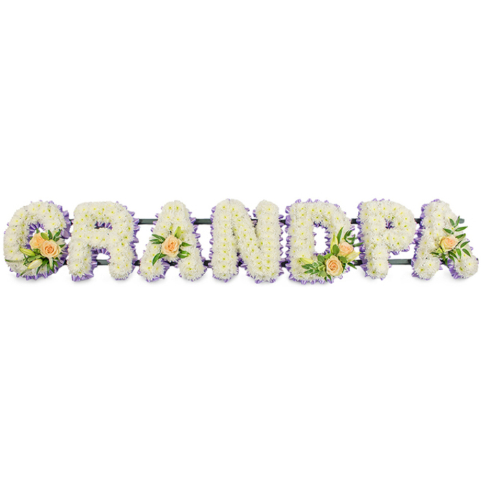 A carefully composed funeral tribute spells out the word GRANDPA in large floral letters, arranged in a straight, confident line on a slim, dark support bar. The design looks as though it could be set at the front of a service in Yiewsley, perhaps at a local church or crematorium, where family and friends come together in remembrance. Each of the seven uppercase letters is tightly massed with pure white chrysanthemums or similar small, button-like flowers, so close together that no gaps show through. Their soft, rounded petals create a plush, almost quilted texture on the surface of every letter, conveying innocence, peace and a sense of gentle farewell. Encircling each letter is a narrow halo of pale lilac blooms, tracing the outline of the G, R, A, N, D, P and finishing A with a subtle wash of colour that separates the forms from the clean white background. The lavender edging is not overpowering; instead it gives definition and a quiet, almost spiritual glow. At key points, peach roses are tucked into the lettering: a generous cluster with a few white lily buds and glossy foliage at the base of the G, smaller accents on the first A and the last A, and a balanced grouping on the D. These roses, with their soft apricot-peach tones, bring warmth and the suggestion of cherished, personal memories. The plain, bright background focuses the eye entirely on the flowers, highlighting the skill of the florist and the freshness of the blooms. This personalised GRANDPA arrangement offers a thoughtful, elegant way for a Yiewsley family to say goodbye, combining local craftsmanship with tender, timeless sympathy flowers.