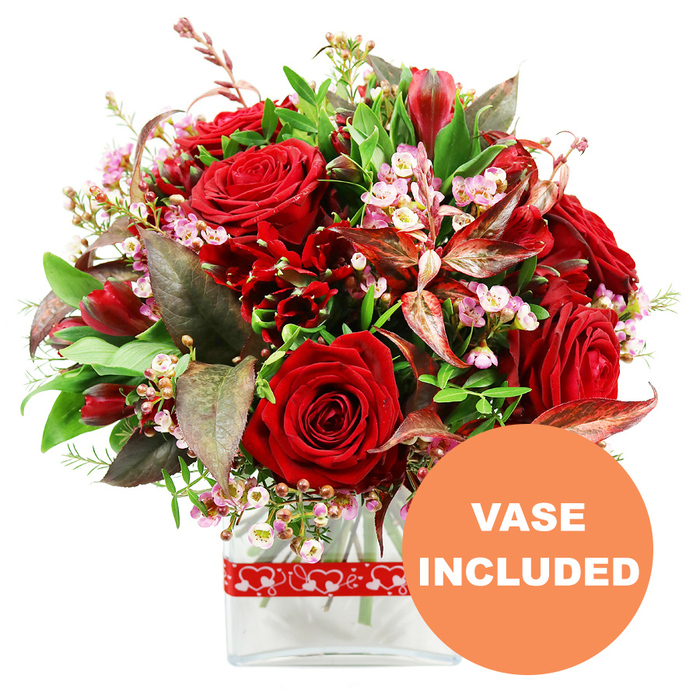 A sumptuous floral display brimming with romantic reds and gentle blush tones stands out dramatically against a pure white background, as if illuminated by soft studio light. At the focal point are several large red roses, their petals thick and velvety, some curled tightly in youthful buds and others open wide, revealing intricate inner whorls that invite the eye to linger. These roses are framed by smaller, equally rich crimson blooms with multiple petals, resembling alstroemeria or similar flowers, whose flared shapes add movement and depth to the arrangement. Interlaced among these vivid reds are delicate sprays of tiny pale pink and white blossoms, like waxflower or limonium, their slender stems and clustered heads softening the composition with a lacy, cloud-like effect. A generous border of greenery surrounds the flowers: glossy green leaves sit alongside lighter, more feathery sprigs, evoking the freshness of a walk through the green spaces around Yiewsley, such as nearby parks and canal-side paths. Striking burgundy and reddish-brown foliage, some subtly variegated, is tucked in thoughtfully, adding shadows, warmth, and a sophisticated contrast that makes the reds appear even more intense. The blooms are arranged in a sleek, transparent glass vase with a rectangular silhouette and gently softened corners, allowing the viewer to glimpse the stems and water within, emphasising the bouquet's freshness as if just prepared in a local Yiewsley flower shop. Around the lower part of the vase, a red ribbon patterned with continuous white hearts wraps neatly, reinforcing the bouquet's purpose as a gesture of love-perfect for anniversaries, Valentine's Day, or an intimate celebration at home. In the bottom right of the image, a bold orange circular overlay clearly announces VASE INCLUDED in sharp white capital letters, underscoring that this romantic, gift-ready piece arrives complete and ready to place on any table or windowsill.