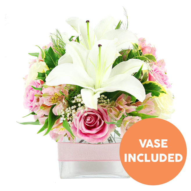 Set against a pure white background, this thoughtfully arranged bouquet presents a graceful mix of white Oriental lilies, pink roses, and airy filler flowers, all nestled in a modern clear glass vase that would sit beautifully on a Yiewsley living room sideboard. In the centre, two impressive white lilies open like trumpets, their wide petals curving outward and catching the light, while dark reddish-brown stamens reach from the centre, adding depth and a touch of drama. Surrounding them, an abundance of soft pink roses fills the middle of the design; some are still in bud, petals wrapped closely like satin ribbons, while others are fully open, revealing their spiralling hearts and giving the arrangement a romantic, storybook quality. Intermixed with the roses are pastel pink and white alstroemeria, their slender petals lightly flecked with yellow, which introduce both pattern and a gentle, almost painterly feel. Small clusters of white baby's breath appear throughout, creating delicate, cloud-like pockets that soften the transitions between the larger flowers. The entire bouquet is framed by rich, glossy greenery and lighter, feathery foliage, as if it were freshly gathered from gardens near the high street in Yiewsley on a calm morning. All of this beauty is carefully composed within a low, square glass vase, its transparent sides revealing the stems and water, and a pale pink satin ribbon tied smoothly around its middle adds a tactile, decorative finish. In the bottom right corner, a bright circular orange graphic with bold white capital letters reads VASE INCLUDED, clearly indicating that this floral arrangement is complete and ready for display, perfect for gifting, celebrating a special milestone, or simply bringing a sense of serene elegance into a Yiewsley home.