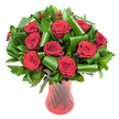A sumptuous, rounded bouquet of a dozen or more deep crimson roses occupies the frame, each bloom opened fully to reveal velvety, tightly spiralled petals whose rich red reads almost wine-dark under even, soft lighting. The roses are clustered close together to create a dense, luxurious crown, their satiny surfaces catching tiny highlights that emphasize petal texture and depth. Surrounding and cradling the heads is a generous sweep of glossy dark green foliage: slim, flat leaves that form a verdant backdrop and broader, gleaming emerald leaves that have been expertly rolled and curved into elegant cylindrical loops. Those folded leaves add an architectural rhythm to the composition, their smooth, reflective surfaces contrasting with the matte softness of the roses. The stems are gathered and displayed in a tall, slender translucent ruby-red glass vase whose subtle ribbed or twisted pattern throws thin vertical lines of light and shadow, echoing the roses' vertical stems and amplifying the arrangement's passionate palette. Set against a pure white background, the crimson and green appear exceptionally vivid, isolated from distraction, and presented with gallery-like clarity. As a working London florist in Yiewsley I find this combination reads as classic romance with a modern twist: formal enough for an anniversary or Valentine's centrepiece, intimate enough for a personal declaration. The scent one imagines is heady and floral, the petals soft to the touch, and the overall silhouette communicates considered craftsmanship and freshness-ideal for a heartfelt gift delivered locally around Yiewsley, West Drayton and neighbouring Stockley Park.
