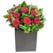 A modern study in romance: a compact cluster of rich, velvety red roses sits like a small sculpture within a matte black cube, each bloom opening in soft, generous spirals that catch the light and deepen the crimson. Broad, glossy green leaves are carefully curled and woven around the roses, their lacquered surfaces forming rhythmic waves that push and pull the eye through the composition. The square vessel-a box-shaped, matt-finished container-grounds the arrangement with a clean, urban silhouette that contrasts with the sensual softness of the petals. Studio lighting is steady and clear, skimming the tops of leaves and throwing gentle highlights across the roses so the crimson seems almost velour to the touch; the foliage's edges shine with a cool, almost tropical crispness. Close up, the bouquet suggests a delicate scent: the classic sweetness of rose tempered by the bright, green notes of fresh leaves and a whisper of cut-stem freshness. As a florist in Yiewsley I picture this arrangement on a low coffee table in a modern flat near the High Street or brightening a reception desk by the Grand Union Canal towpath-its presence both decisive and intimate. It's the kind of bouquet chosen for a quietly powerful declaration: an anniversary that asks to be remembered, a thoughtful thank you, or a minimalist wedding table accent. Texturally and emotionally, it balances drama and restraint, offering a composed statement that feels both immediately present and thoughtfully crafted for the neighbourhood's contemporary homes and small celebrations.