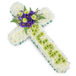 A dignified floral cross is captured from a slightly angled view, laid out on a clean white surface so that its colours and textures stand out clearly. The main structure is built from masses of pure white spray chrysanthemums, closely packed to create a thick, cushioned layer that feels soft and consoling, like a blanket of petals. Around the very edge of the cross, a bright, fresh green border made from ribbon or carefully tucked foliage traces the outline, giving a sharp, defined finish to the arms and stem and echoing the greenery found in nearby parks around Yiewsley. Down the centre of the vertical arm, a panel of lime green button chrysanthemums adds a refreshing note of colour and a slightly raised texture. Delicate, almost thread-like wires in pale green or silver are looped and crossed over this strip, forming a gentle lattice that lends a touch of intricate craftsmanship. At the heart of the cross, where the two arms intersect, a cluster of deep purple lisianthus blooms forms a rich focal point, their velvety petals folding in layers of royal colour. Nestled among them are lime green button mums and sprigs of dark ferns and foliage, which fan out subtly to create depth and contrast against the white chrysanthemum base. The lighting is soft and even, enhancing the calm mood of the design. This cross communicates remembrance, faith, and tender farewell, the kind of tribute families in Yiewsley might choose to accompany a final goodbye at a funeral or memorial service.